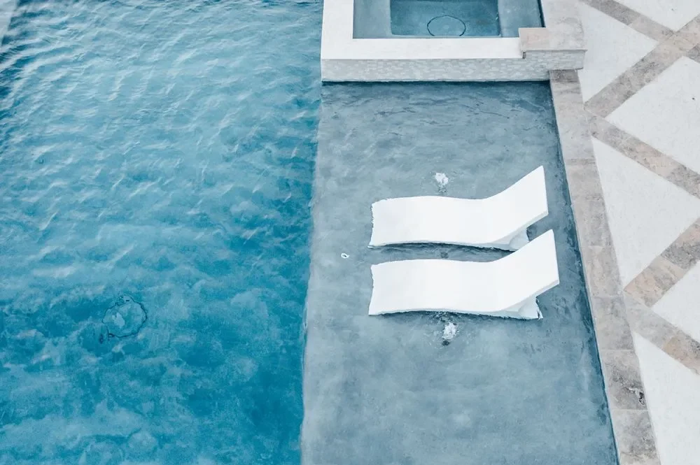 Aerial view of two white lounge chairs partially submerged in a clear blue swimming pool surrounded by a textured stone deck