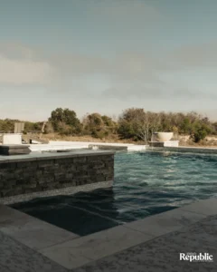 A tranquil poolside scene features a sleek water feature, lounge chairs, and surrounding greenery under a soft, cloudy sky